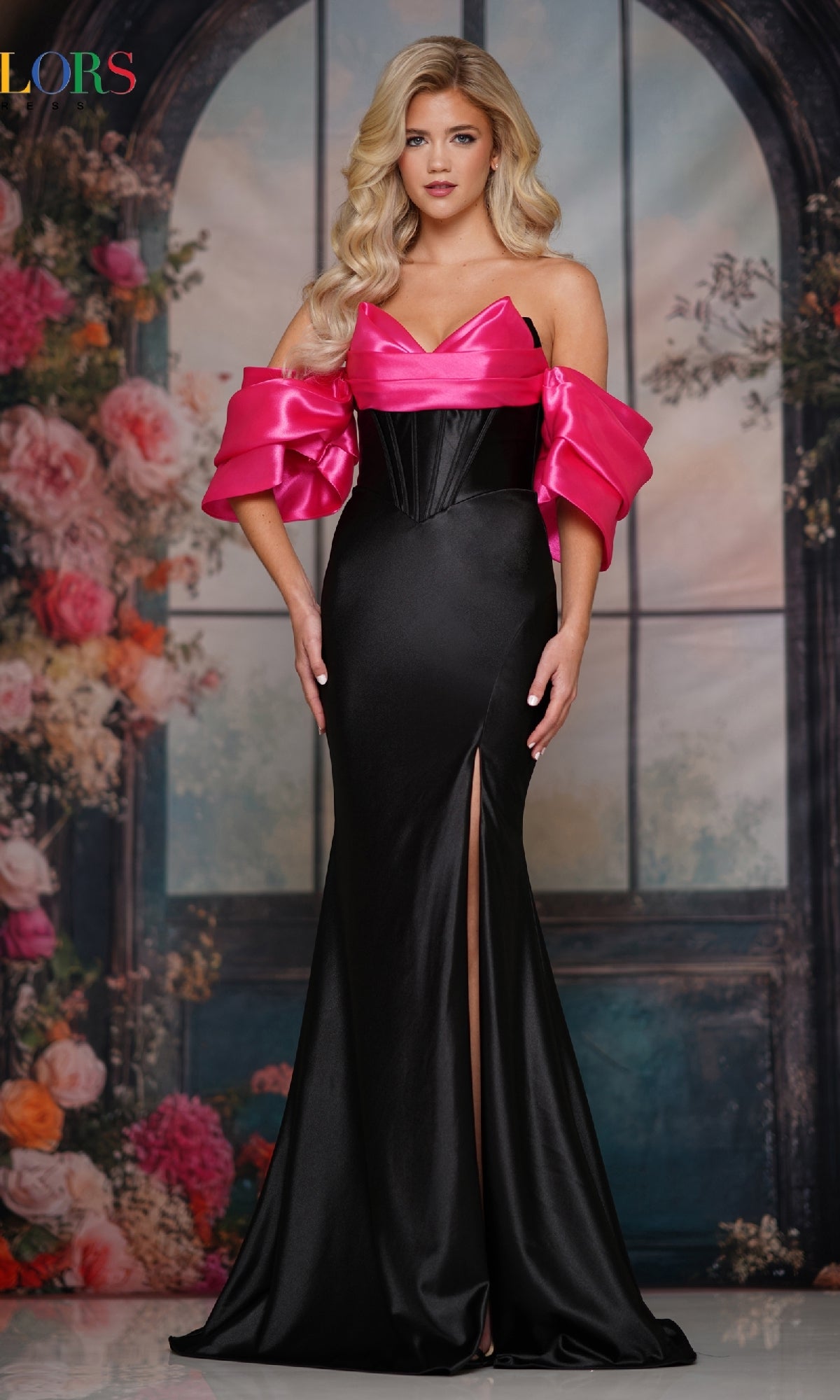 A woman poses before a floral backdrop in the Strapless Long Black Prom Dress: Colors Dress 3878, featuring a black and pink corset-style bodice, sweetheart neckline, and front leg slit.